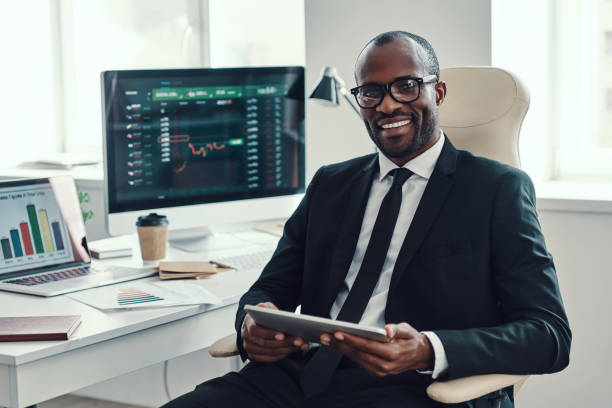 istockphoto-1194668220-612x612 Concentrated young African man in formalwear using modern technologies and smiling while working in the office